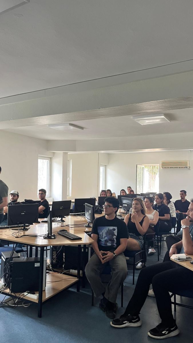 In a bright computer laboratory, approximately twenty students are seated at semicircular desks. On the desks are black computer monitors, keyboards, and mice. Near the camera, an upside-down computer monitor rests on a desk.  At the front of the classroom, in front of a whiteboard and a projection screen, an instructor is giving a lecture. A formal meeting-style presentation is projected onto the screen.  The students are attentively looking forward; some are resting their elbows on the desks, while others are slightly turned toward the instructor.  The classroom has white walls and a high ceiling. On the front wall, there is a clock and a framed portrait of Mustafa Kemal Atatürk. The overall atmosphere is calm and focused on learning.